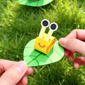 Puede incluir: Una manualidad de caracol hecha de papel amarillo con una concha en espiral, dos tallos verticales con ojos circulares blancos y negros, y una boca roja sonriente, todo montado sobre una hoja de papel verde con venas dibujadas.