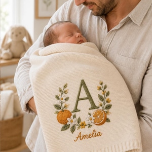Puede incluir: Una manta de punto para bebé de color crema con un diseño bordado de una letra A grande en verde rodeada de naranjas, hojas verdes y pequeñas flores blancas. El nombre Amelia está bordado en letra cursiva naranja debajo del diseño.