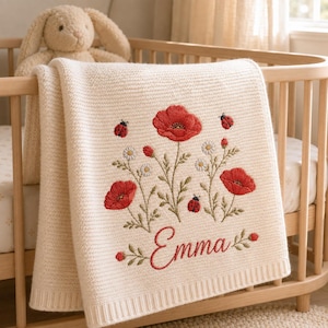 Könnte beinhalten: Eine cremefarbene Strickdecke für Babys mit einer Stickerei aus roten Mohnblumen, kleinen weißen Gänseblümchen und Marienkäfern. Der Name Emma ist in roter Schreibschrift unten in der Mitte eingestickt. Ein beigefarbener Plüschhase steht dahinter.