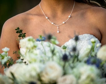 May include: A bride wearing a white strapless wedding dress with a pearl necklace. She is holding a bouquet of white flowers with green leaves and blue accents.
