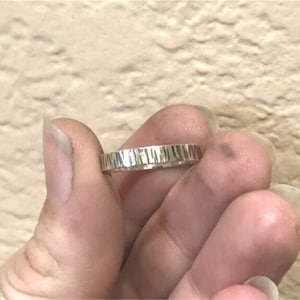 May include: A silver ring with a textured, hammered finish. The ring is shown on a finger.