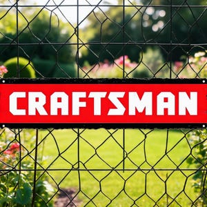 May include: A rectangular metal sign with a bright red background and a thick black border. The word CRAFTSMAN is printed in large, bold, white capital letters across the center. The sign features small circular holes at each corner for mounting.