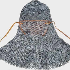 Puede incluir: Una cofia de cota de malla de color plateado con una cubierta redondeada para la cabeza que cae sobre los hombros. La pieza incluye una máscara facial de cuero marrón en la parte delantera, asegurada con finas tiras de cuero marrón.