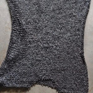 May include: A rectangular piece of chainmail armor constructed from numerous small, interlocking dark grey metal rings. The top edge features a scalloped, jagged pattern, while the overall structure displays a dense, uniform mesh texture.
