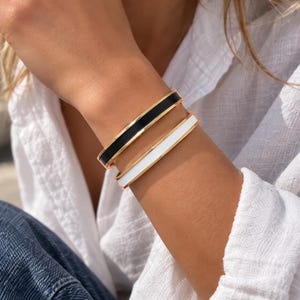 May include: Two slim bangle bracelets worn on the wrist. One bracelet features a polished gold-tone metal band with a central inlay of solid black enamel. The second bracelet features a matching gold-tone metal band with a central white enamel inlay.