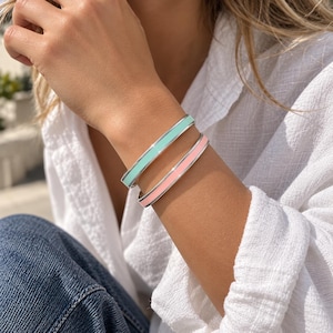 May include: Two slim metal bangle bracelets featuring smooth enamel inlay. One bracelet is finished in a soft mint green colour, while the other is finished in a pale pastel pink. Both bangles have a polished silver-tone metal border.