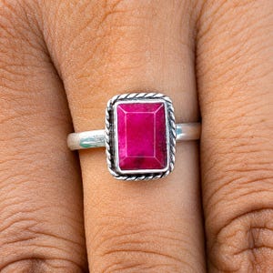 May include: A silver ring featuring a rectangular, faceted, deep pink gemstone set in a twisted rope-style metal border. The band is smooth and polished, showcasing a clean, metallic finish that complements the vibrant, saturated color of the stone.