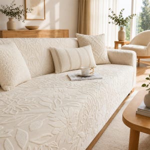 May include: A cream-coloured sofa cover featuring a raised floral pattern, paired with a rectangular lumbar pillow with vertical stripes and a square textured throw pillow. The fabric has a soft, embossed botanical design covering the entire surface.