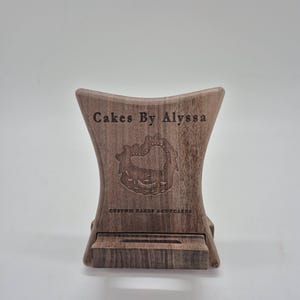 Peut inclure: Un support de téléphone en bois brun avec une silhouette incurvée, présentant le texte gravé "Cakes By Alyssa" au-dessus d'un motif en forme de cœur et les mots "CUSTOM CAKES & CUPCAKES" en bas, avec une fente horizontale pour l'appareil.