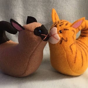 May include: Two felt cat toys, one brown and one orange, are facing each other with their heads touching. The brown cat has black ears and a black tail. The orange cat has a curled tail and orange stripes.