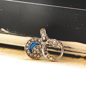 May include: A silver-toned metal charm featuring two circular components. One circle contains a blue crescent moon surrounded by clear crystals. The second circle is an open ring decorated with small star shapes and clear crystals.