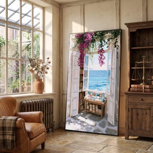 Puede incluir: Un gran cuadro rectangular enmarcado que representa una ventana abierta con vistas a un mar azul brillante. La escena muestra contraventanas blancas, flores de buganvilla de color rosa intenso y una mesa de madera llena de libros.