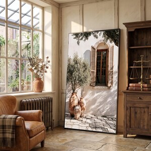 Könnte beinhalten: Ein großer rechteckiger Standspiegel mit einem aufgedruckten Bild einer mediterranen Gebäudefassade, inklusive Fenster, Holzfensterläden, drei Terrakottatöpfen mit kleinen Bäumen und einem gepflasterten Boden in einer hellen Umgebung.