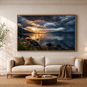 Op de afbeelding: Een grote landschapsmuurprint in een houten lijst met een zonsondergang boven een meer met donkere, dramatische wolken, rotsachtige oevers en goudkleurig licht dat reflecteert op het water, weergegeven boven een beige bank in een woonkamer.