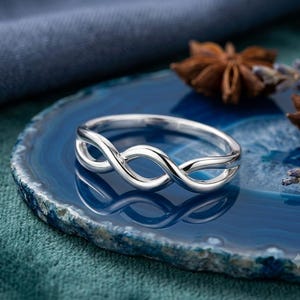 Puede incluir: Un anillo de plata pulida con un diseño de bucle infinito continuo. La banda está construida con dos hilos de metal entrelazados que crean un patrón simétrico y calado, reflejando la luz en su superficie lisa y curva.