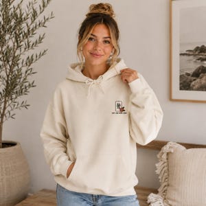 Könnte beinhalten: Ein cremefarbener Kapuzenpullover mit einer Kängurutasche auf der Vorderseite und einer kleinen Grafik auf der linken Brust. Die Grafik zeigt eine rechteckige Spielkarte mit einem schwarzen Schriftzeichen, einer kleinen roten Blume und dem Text "JUST ONE MORE HAND".