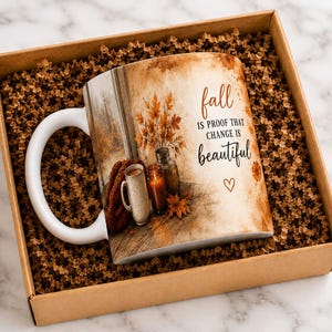 May include: A white ceramic mug featuring a printed autumn scene with a candle, a beverage, and fall foliage. The text reads: fall IS PROOF THAT CHANGE IS beautiful, accompanied by a small heart icon. The mug is nestled in a brown cardboard gift box.