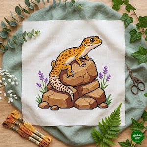 May include: A cross-stitch embroidery on white fabric featuring a yellow and brown spotted leopard gecko perched on a stack of brown rocks, flanked by small purple lavender sprigs and green grass blades, with embroidery floss and scissors nearby.