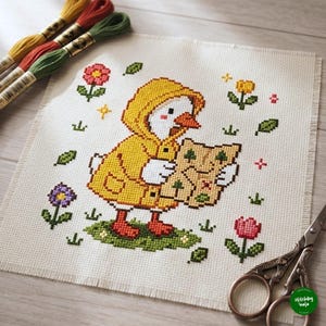 May include: A cross-stitch embroidery on white fabric featuring a white duckling wearing a yellow raincoat and holding a map. The design includes small colorful flowers, green leaves, and grass. Three skeins of embroidery floss and scissors are nearby.