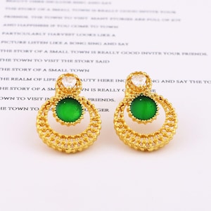 May include: A pair of gold-toned earrings featuring a circular filigree frame, a central round green stone, and a small clear heart-shaped stone at the top. The intricate metalwork creates a delicate, openwork pattern around the green center.