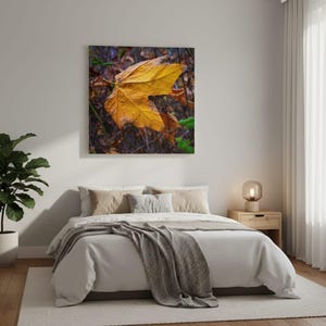 May include: A square wall art print featuring a large, vibrant yellow maple leaf centered against a dark, textured background of fallen leaves and thin branches, with small patches of green foliage visible in the lower right corner.