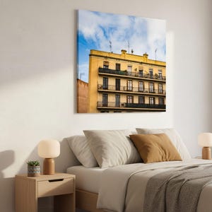 May include: A square wall art print featuring a yellow building with multiple balconies and dark window frames against a bright blue sky with white clouds. The building has a distinct architectural style with several levels of iron railings.