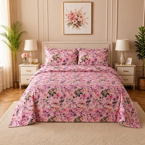 May include: A bed featuring a comforter and two matching pillow shams with a pink, purple, and white floral pattern. The bedding set is displayed on a bed frame with a headboard, flanked by two white nightstands with lamps and a framed floral print.