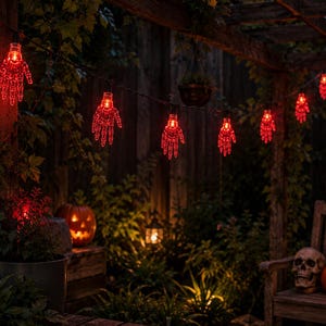 Peut inclure: Une guirlande lumineuse décorative composée de caches en forme de mains de squelette rouges et translucides. Les lumières sont suspendues à un fil sombre, projetant une lueur rouge vive dans l'espace extérieur, créant un effet visuel marqué.
