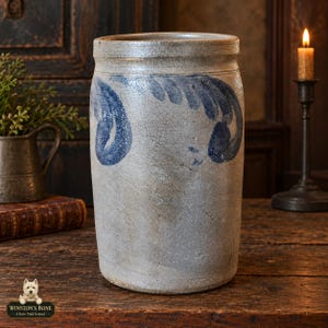 May include: A cylindrical stoneware crock with a light gray, textured finish and a prominent blue cobalt slip-trailed leaf design around the upper body. The vessel features a thick, slightly flared rim and a flat base, showcasing a weathered appearance.