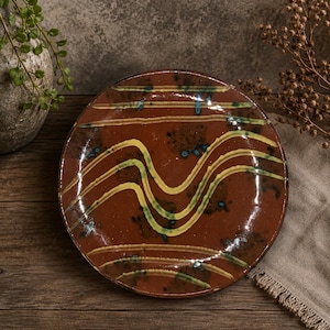 May include: A round ceramic plate with a glossy dark brown glaze featuring hand-painted wavy yellow lines across the center and straight horizontal yellow stripes at the top and bottom, accented by small dark blue and black speckled details.