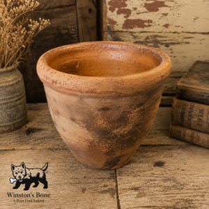 May include: A round, terracotta-colored clay pot with a thick, rounded rim and a weathered, textured surface showing variations in brown and tan tones. The vessel has a wide opening and a tapered base, displaying a rustic, aged appearance.