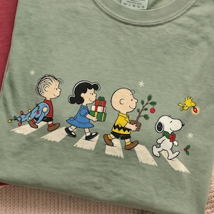 May include: Three crew neck t-shirts in cream, maroon, and sage green. The sage green shirt features a graphic print of the Peanuts characters walking in a line, carrying Christmas items, with the Comfort Colors brand label visible at the neck.