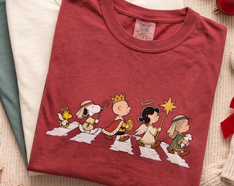 May include: Three crew neck t-shirts in dark green, cream, and maroon. The maroon shirt features a graphic of Peanuts characters dressed as a nativity scene walking on a crosswalk. The shirt label reads Comfort Colors with size and care details.