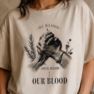 以下が含まれることがあります： ベージュの半袖Tシャツで、2つの手が握り合っている黒いグラフィックが描かれています。テキストには「MY BLOOD」「YOUR BLOOD」「OUR BLOOD」とあり、小さな三日月、ローズマリーの小枝、ベリーのイラストが添えられています。