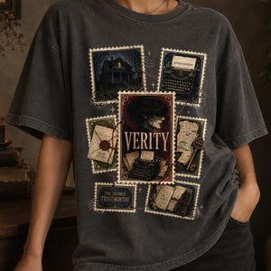 May include: A charcoal gray short-sleeve t-shirt featuring a graphic print of multiple postage stamp-style illustrations. The central design includes the word VERITY, a typewriter, a sealed envelope, a key, and a house, all in a dark aesthetic.