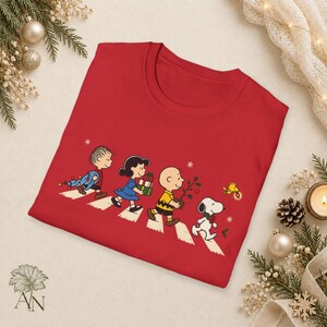 May include: A red t-shirt featuring a graphic of Linus, Lucy, Charlie Brown, Snoopy, and Woodstock walking in a line across a crosswalk. The characters are carrying holiday items like gifts, a small tree, and string lights, with snowflakes.