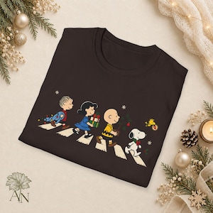 May include: A dark brown crew neck t-shirt featuring a graphic of Linus, Lucy, Charlie Brown, Snoopy, and Woodstock walking across a crosswalk. The characters are carrying Christmas items including gifts, a small tree, and holiday lights.