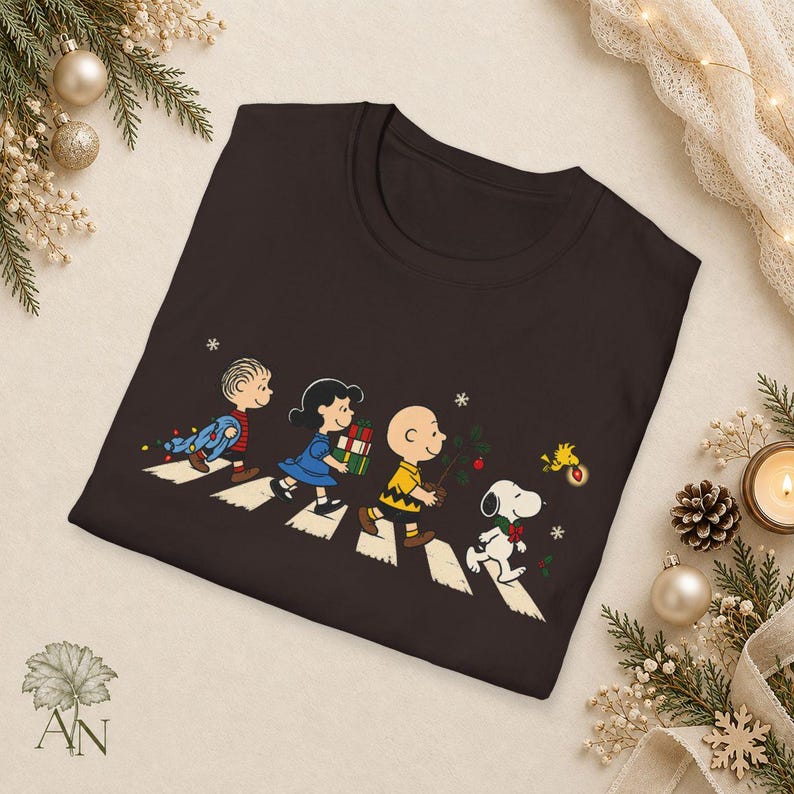 May include: A dark brown crew neck t-shirt featuring a graphic of Linus, Lucy, Charlie Brown, Snoopy, and Woodstock walking across a crosswalk. The characters are carrying Christmas items including gifts, a small tree, and holiday lights.