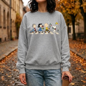 May include: A heather gray crewneck sweatshirt featuring a graphic print of the Peanuts characters walking in a line across a crosswalk. The design includes Linus, Lucy, Charlie Brown, and Snoopy carrying holiday items like gifts and a small tree.