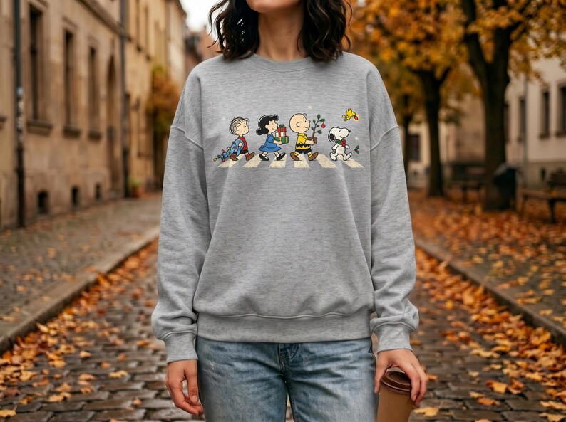 May include: A heather gray crewneck sweatshirt featuring a graphic print of the Peanuts characters walking in a line across a crosswalk. The design includes Linus, Lucy, Charlie Brown, and Snoopy carrying holiday items like gifts and a small tree.