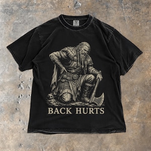 May include: A black short-sleeve t-shirt featuring a graphic print of a bearded warrior in fur-trimmed armor kneeling while holding an axe. The warrior has one hand on their lower back. The text BACK HURTS is printed in a light beige font below.