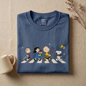 May include: A navy blue crew neck t-shirt featuring a graphic of the Peanuts characters walking across a crosswalk. The design includes Linus, Lucy, Charlie Brown, Snoopy, and Woodstock with holiday items. The inner label reads Comfort Colors.