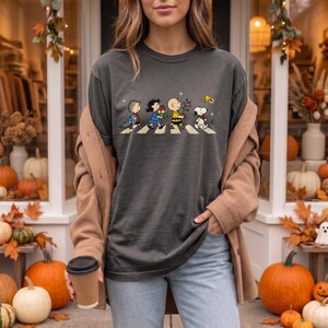May include: A charcoal gray t-shirt featuring a graphic of the Peanuts characters walking in a line across a crosswalk. The design includes Linus, Lucy, Charlie Brown, Snoopy, and Woodstock, with small holiday elements like gifts and a tree.
