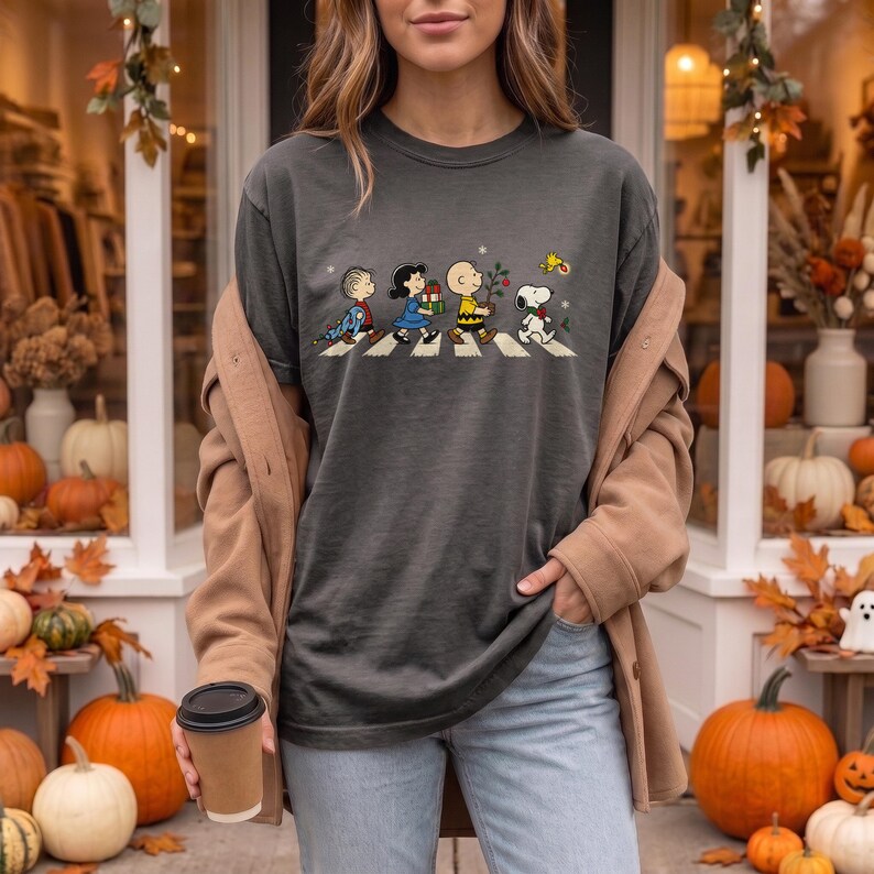 May include: A charcoal gray t-shirt featuring a graphic of the Peanuts characters walking in a line across a crosswalk. The design includes Linus, Lucy, Charlie Brown, Snoopy, and Woodstock, with small holiday elements like gifts and a tree.