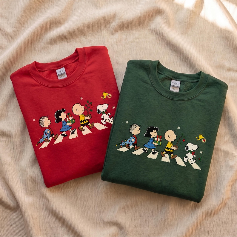 May include: A red crewneck sweatshirt and a dark green crewneck sweatshirt, both featuring a graphic print of the Peanuts characters walking in a line across a crosswalk, carrying Christmas gifts, a small tree, and holiday lights.