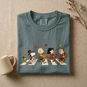May include: A slate blue t-shirt featuring a graphic of the Peanuts characters walking across a crosswalk with autumn leaves. The design includes Snoopy, Woodstock, Charlie Brown, Lucy, and Linus carrying pumpkins and fall foliage items.