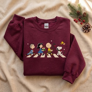 May include: A maroon long-sleeve crewneck sweatshirt featuring a graphic of the Peanuts characters Linus, Lucy, Charlie Brown, Snoopy, and Woodstock walking across a crosswalk, carrying Christmas gifts, a small tree, and holiday decorations.