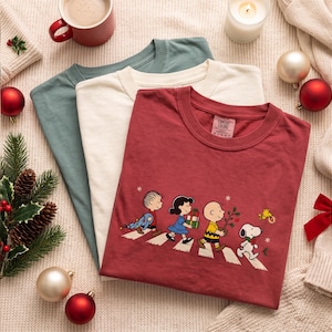 May include: Three crew neck t-shirts in dark red, cream, and muted green. The red shirt features a graphic of Linus, Lucy, Charlie Brown, Snoopy, and Woodstock walking in a line across a crosswalk, carrying Christmas items and a small tree.