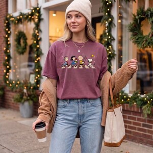 May include: A maroon short-sleeve t-shirt featuring a graphic print of the Peanuts characters walking in a line across a crosswalk, paired with light blue denim jeans, a tan corduroy jacket, a beige knit beanie, and a beige canvas tote bag.