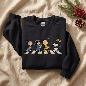 May include: A black long-sleeve sweatshirt featuring a graphic print of the Peanuts characters Linus, Lucy, Charlie Brown, Snoopy, and Woodstock walking across a crosswalk, carrying Christmas gifts and a small tree with snowflakes.